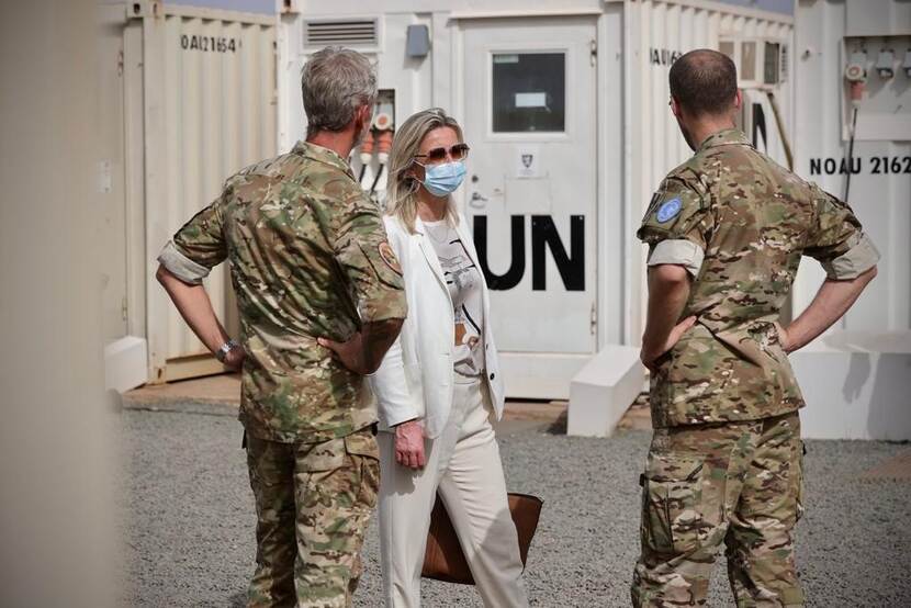 Minister met zonnebril en mondkapje staat tussen 2 militairen. Op achtergrond een barak met letters UN.