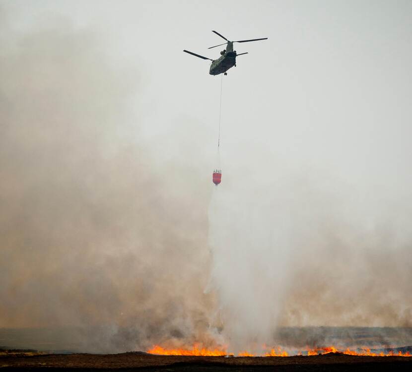 Chinook met bambi bucket vliegt boven landschap in brand.