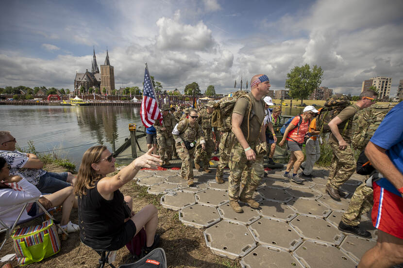 Groep Amerikaanse militairen zijn net de Waal overgestoken tijdens de 4Daagse, een van hen draagt de Amerikaanse vlag. Kerk op de achtergrond.