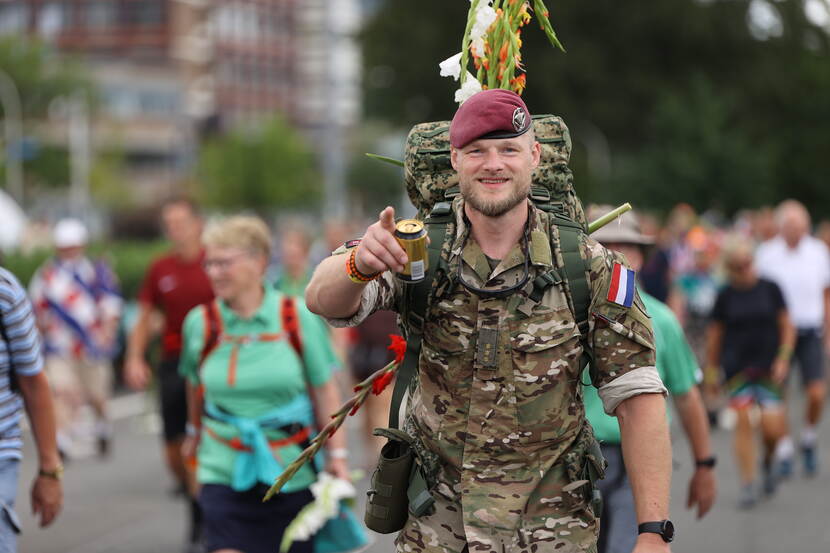 Militair met rode baret bij intocht 4Daagse. Op z'n rug bepakking en een bos gladiolen.