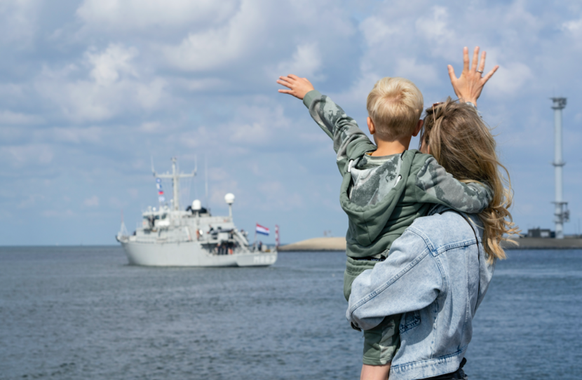 Moeder met kind op de arm zwaaien naar wegvarend marineschip Zr.Ms. Vlaardingen.