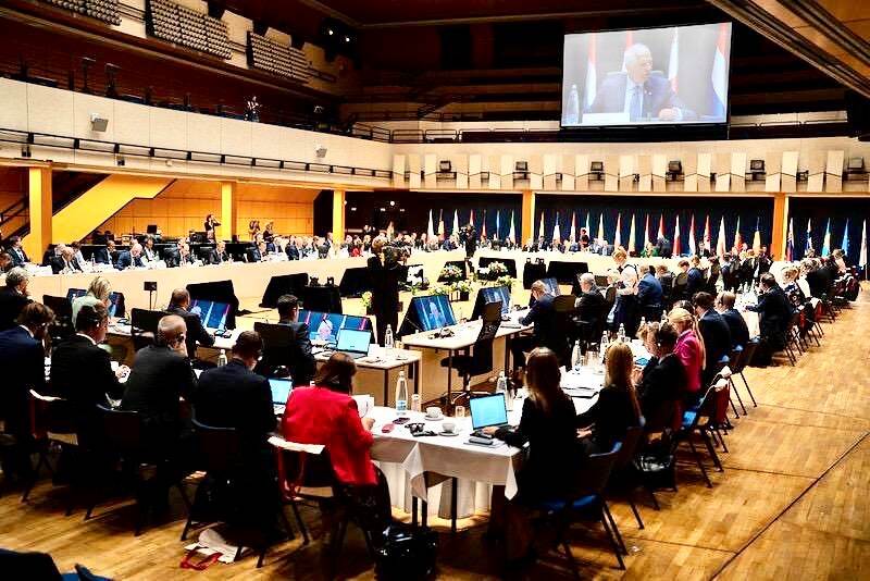 Leden van de informele Raad Buitenlandse Zaken/Defensie aan tafels in een grote zaal.