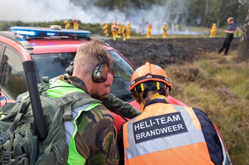 3 mannen in overleg, staand bij een auto. Brandend gebied op de achtergrond.