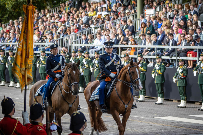 Gouverneur der Residentie te paard inspecteert het militair ceremonieel, publiek op de achtergrond.