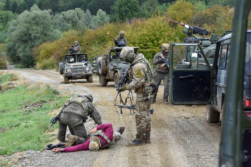 Militairen houden man die op de grond ligt onder schot. Voertuigen in lijn rechts.