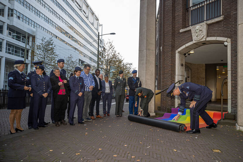 2 militairen rollen regenboogvlag uit bij ingang ministerie. Rijtje militairen kijkt toe.