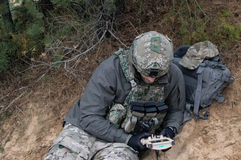 Militair in gevechtspak zit op de grond en kijkt op digitaal scherm.