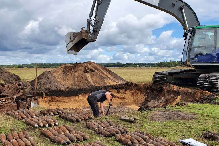 Graafmachine naast een gegraven gat in een weiland, tientallen bommen liggen ernaast.