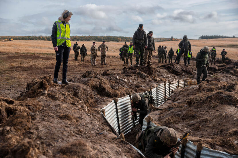 Minister kijkt naar militairen die golfplaten plaatsen in een kuil.