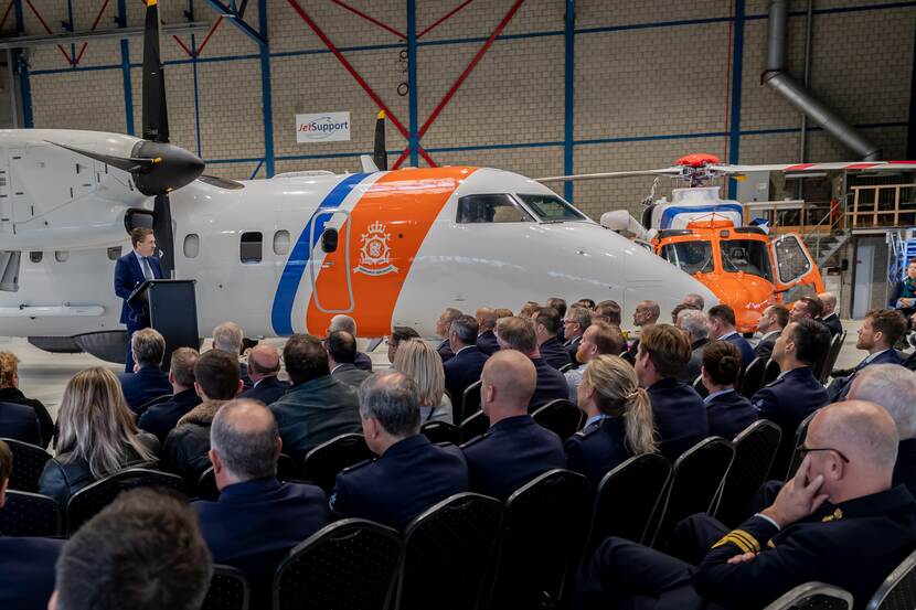 Groep mensen luisteren naar spreker, een nieuw Dash-8-patrouillevliegtuig en AW189 Search & Rescue-helikopter op de achtergrond.