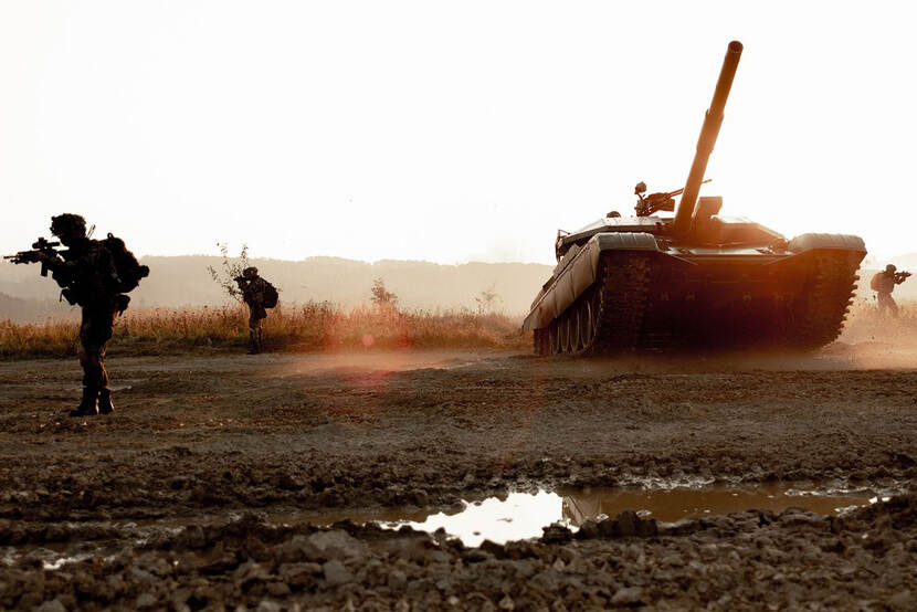 3 militairen met wapen bij een tank, in modderig gebied.