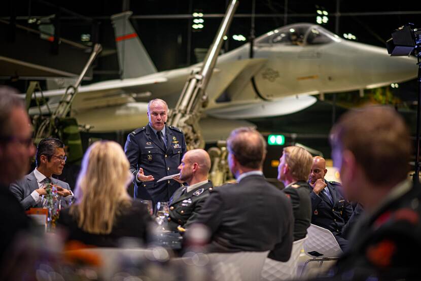 Commandant der Strijdkrachten spreekt voor publiek. Gevechtsvliegtuig staat op de achtergrond.