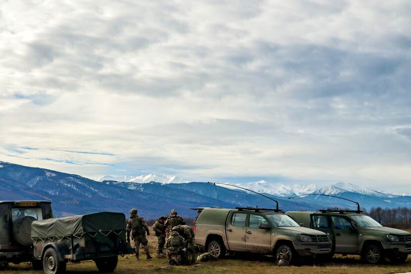 Militairen en voertuigen op voorgrond. Achtergrond: besneeuwde bergen en dik pak witte wolken.