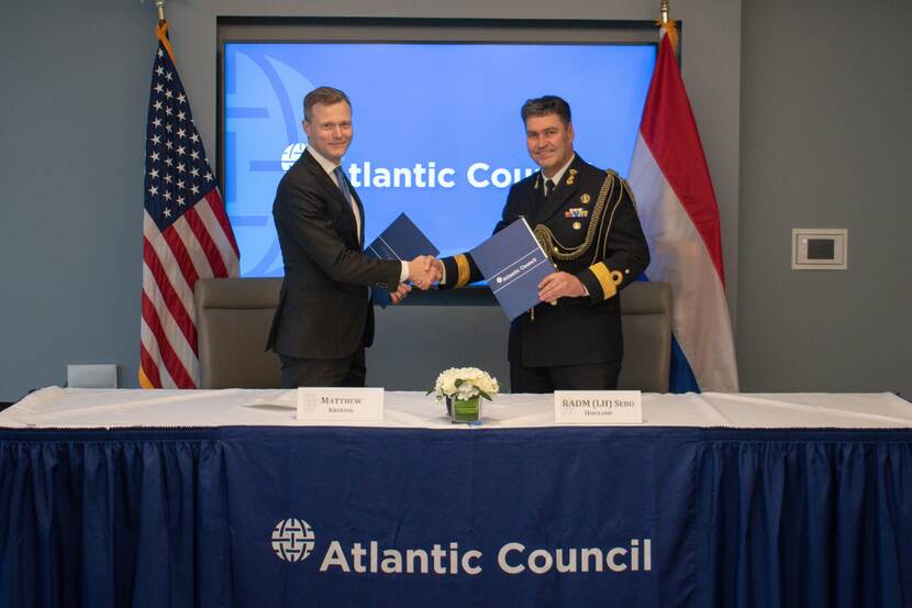 Dr. Matthew Kroenig en commandeur Sebo Hofkamp schudden elkaar de hand voor een beeldscherm met de tekst Atlantic Council. Landsvlaggen aan weerszijden.