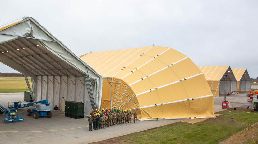 Een luchtfoto van een shelter voor F-35’s, met een groep mensen ervoor.