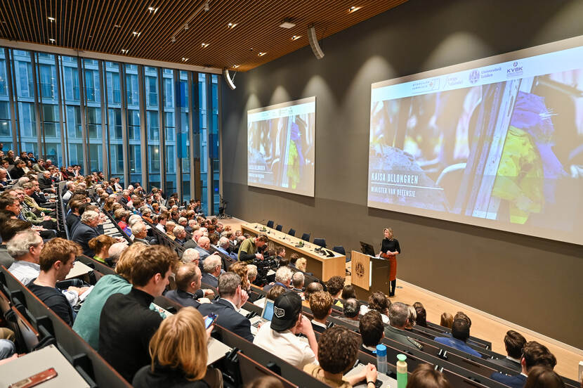 Volle collegezaal met spreker minister Ollogren achter katheder.