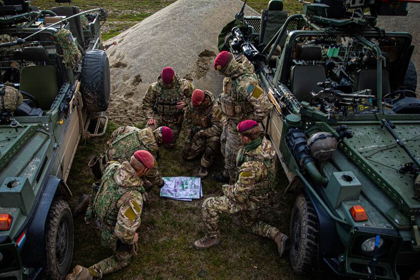 6 militairen van de Luchtmobiele Brigade zitten geknield tussen 2 voertuigen en bestuderen een kaart.