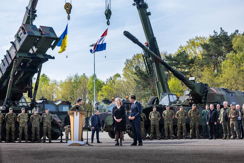 De Oekraïense president Volodymyr Zelensky bedankt Defensiepersoneel. Premier Rutte, minister Ollongren en Defensiepersoneel luisteren. Groot materieel op de achtergrond.