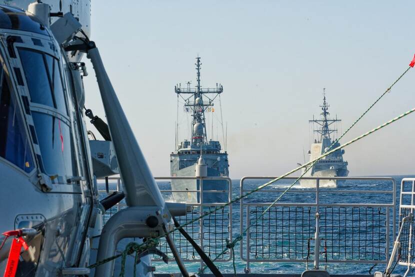 2 fregatten op volle zee, gezien vanaf het dek van een derde schip. Links in beeld een deel van een helikopter.