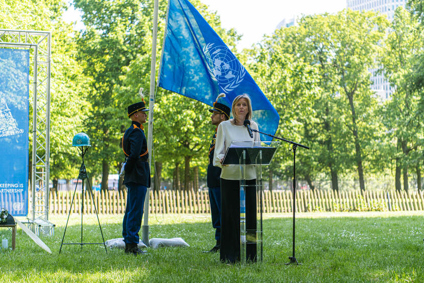 De minister speecht. Achter haar 2 militairen en een blauwe vlag.