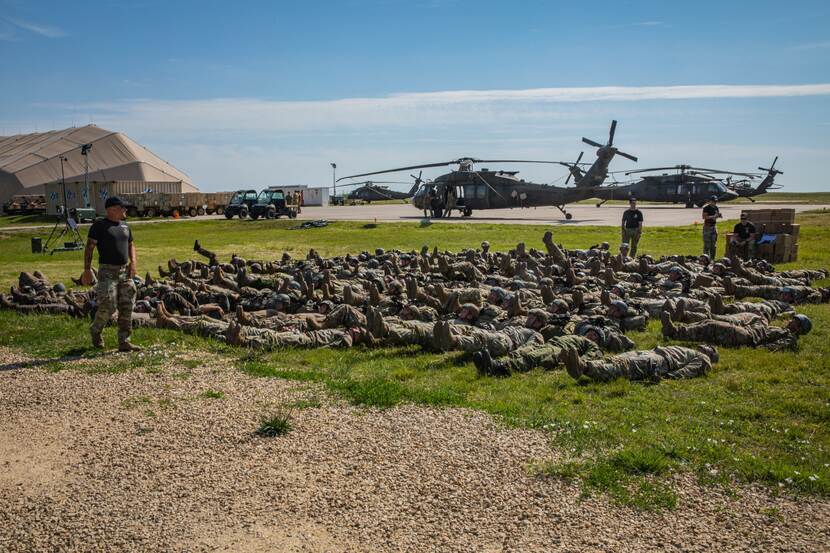 Militairen doen oefeningen op de grond. Op de achtergrond 2 helikopters.