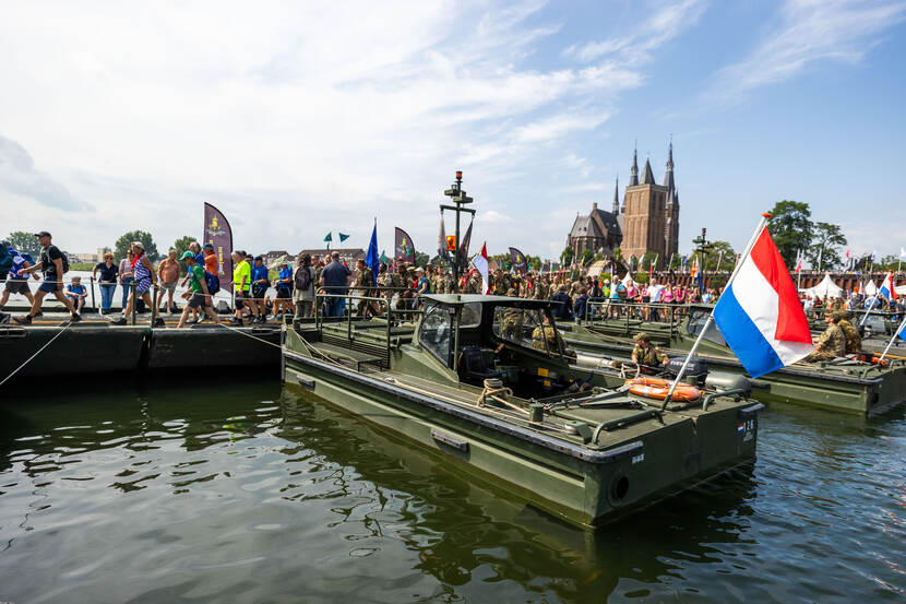 4Daagse wandelaars lopen over een door militairen gelegde pontonbrug. Militaire vaartuigen liggen haaks op de pontonbrug.