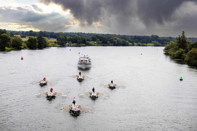 6 sloepen met roeiers varen in V-formatie over een brede rivier onder donkere wolken. Achter hen een groot schip met ondersteuners.