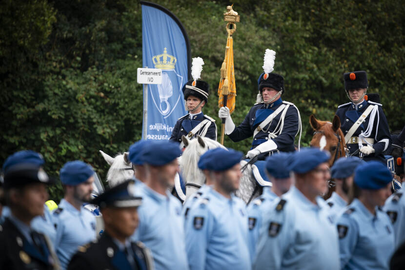 Marechaussees te paard rijden achter rij militairen langs.