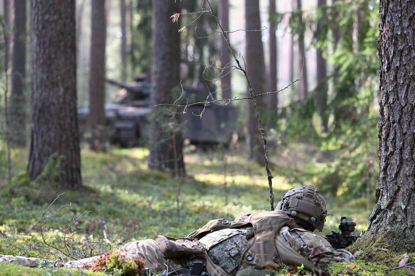 Militair voertuig tussen de bomen. Op de voorgrond ligt een militair met wapen.