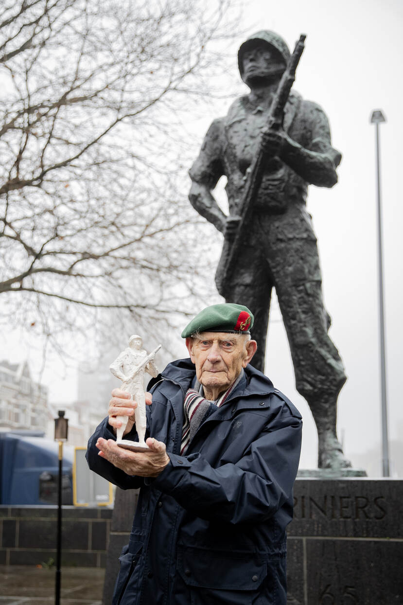 Een veteraan toont een beeldje, met op de achtergrond een standbeeld.