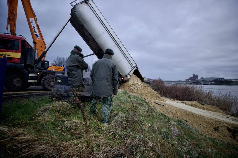 Vrachtwagen lost zand op een dijk. 2 militairen kijken toe.