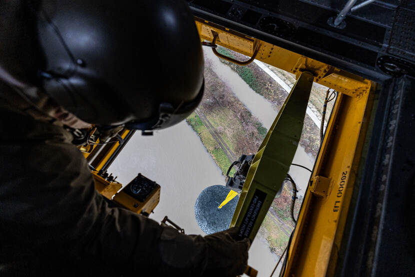 Loadmaster kijkt vanuit Chinook naar lading onder zich, een groot net met zware stenen.