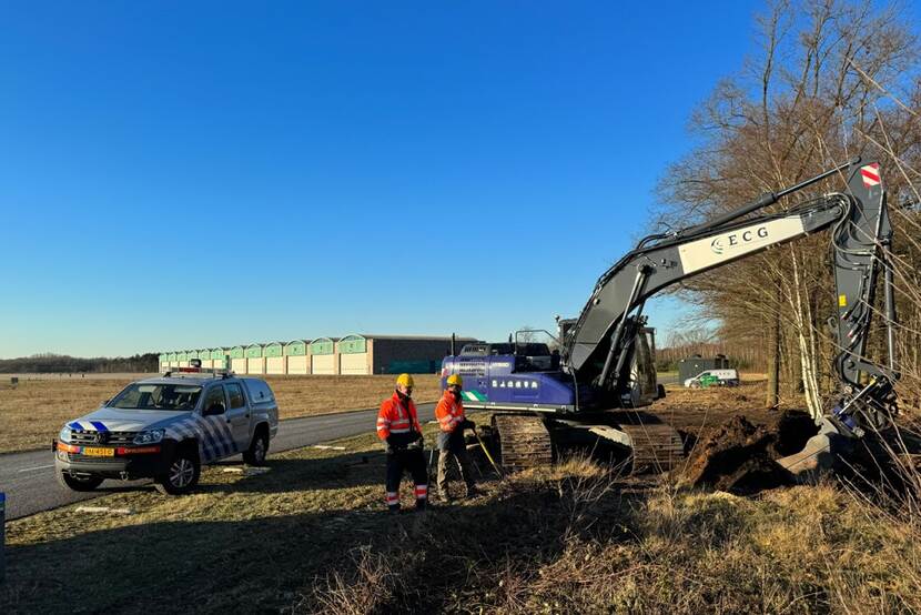 Een graafmachine van de EOD aan de slag in Brabant, loodsen op de achtergrond.