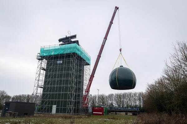 Een mobiele hijskraan hijst een geluidwerende koepel omhoog om deze te plaatsen op het militaire radarstation in het Friese Wier. Het radarstation staat in de steigers.