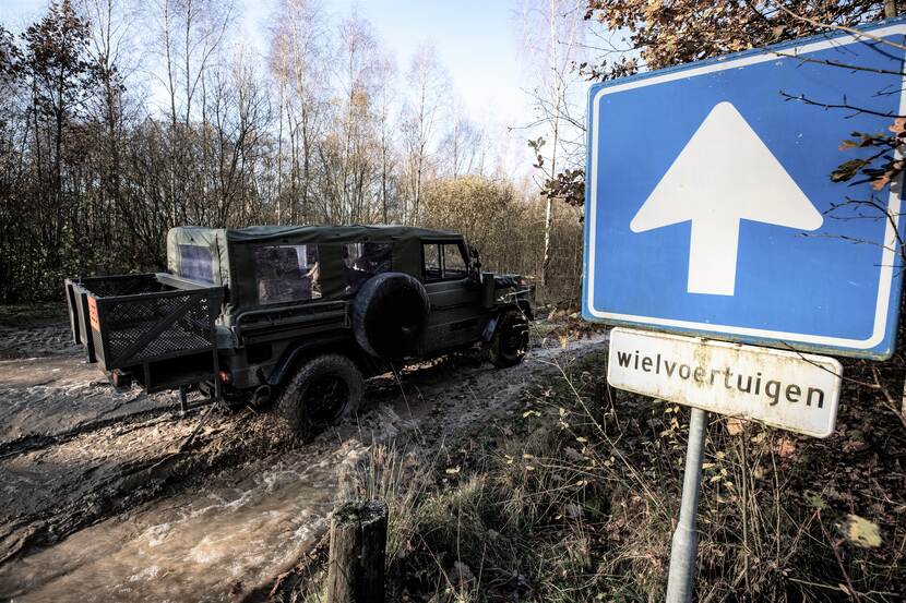 Een jeep rijdt door het bos, langs een verkeersbord met een pijl en de tekst 'wielvoertuigen'.