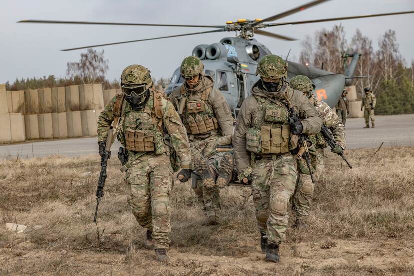 4 militairen dragen gewonde collega op brancard. Op de achtergrond staat een helikopter.