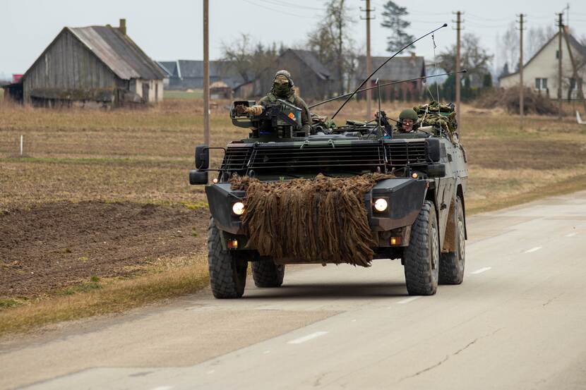 Militair gevechtsvoertuig rijdt op openbare weg tussen paar schuren en huizenrijen.
