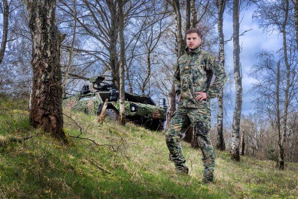 Een mannelijke militair poseert met het nieuwe tenue voor de landmacht in een bos met op de achtergrond een militair voertuig.