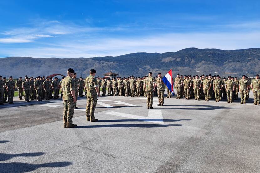 Opgestelde militairen tegen achtergrond van bergen en blauwe lucht. Militair overhandigt Nederlandse vlag.