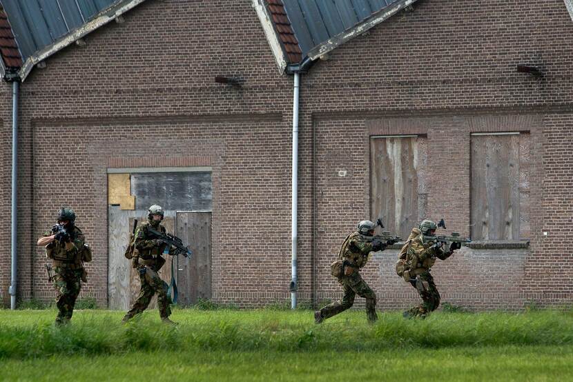4 Commando's rennen en schieten voor een gebouw langs.