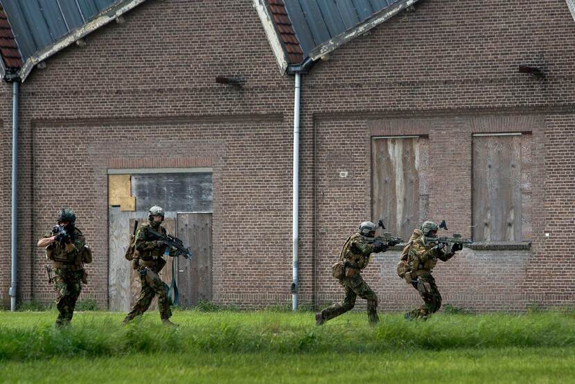 4 Commando's rennen en schieten voor een gebouw langs.