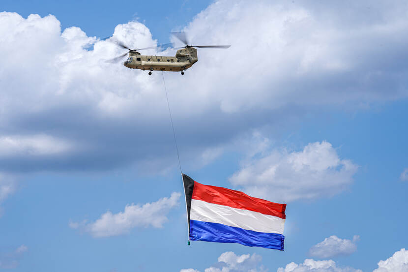 Chinook-transporthelikopter in de lucht met daaronder enorme Nederlandse vlag.