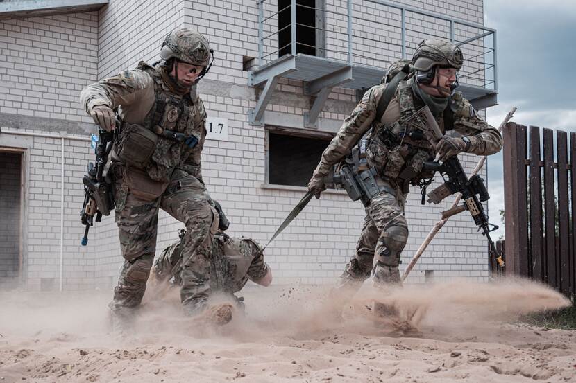 2 militairen snellen pand uit. In hun midden gewonde militair. Opstuivend zand.