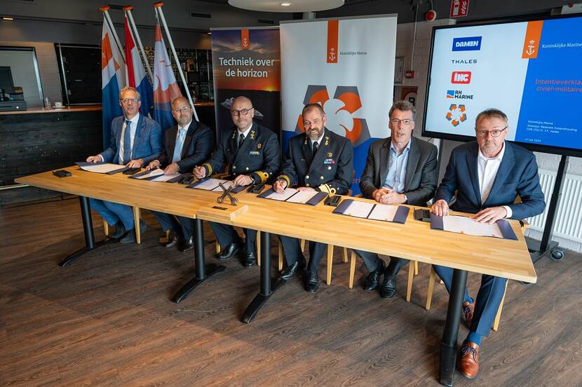 De 6 afgevaardigde mannen van de betrokken partijen zitten aan een tafel met de overeenkomst voor zich.