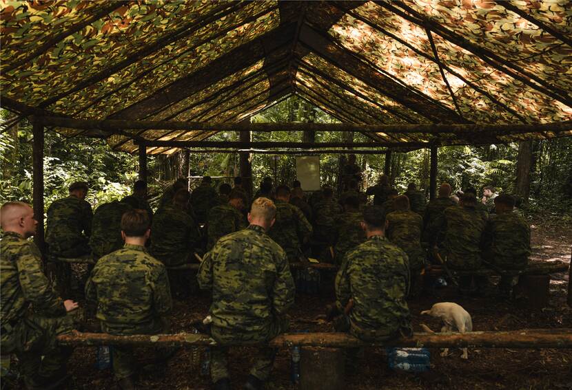 Een groep militairen krijgt les onder een groen net, in de bossige duisternis van de Surinaamse Jungle.