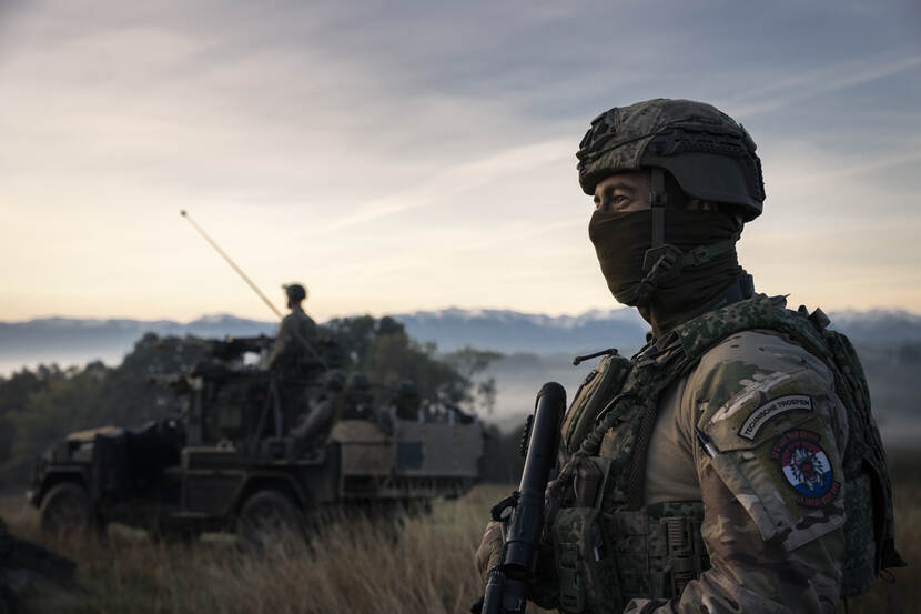 Een militair kijkt voor zich uit. Hij draagt een balaclava. De zon komt net op. Achter hem staat een voertuig.