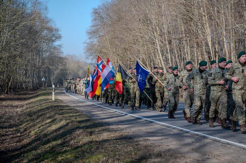 Militairen in lange rij met vlaggen marcheren langs bosrand.