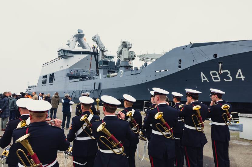 Marineschip ligt langs kade. Op voorgrond militaire muziekkapel.