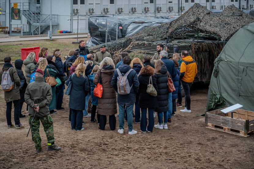 Mensen in burger luisteren naar militair voor de groep. Op de achtergrond militair materieel.