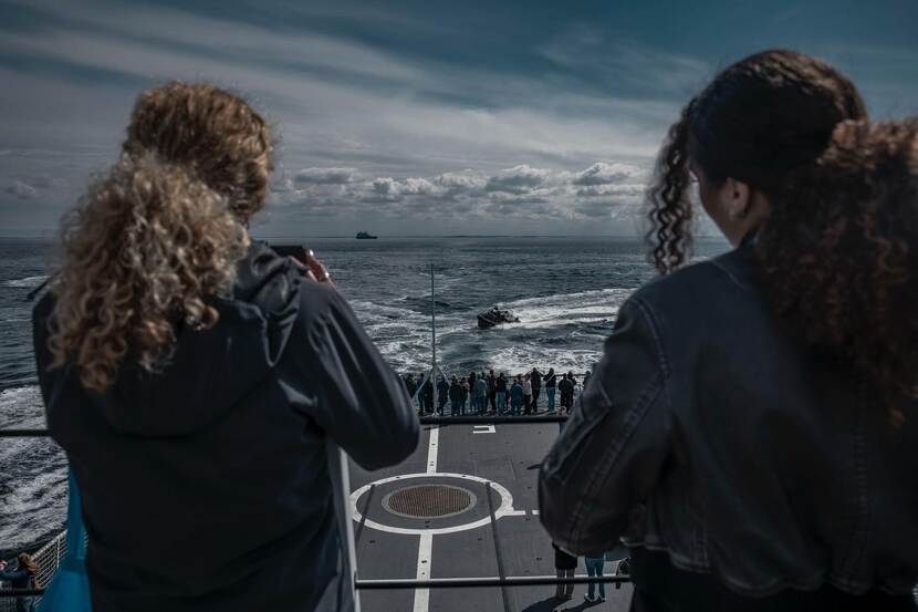 2 meiden op marineschip kijken vanaf boven naar dek beneden. Op zee vaart de snelle motorboot Frisc.